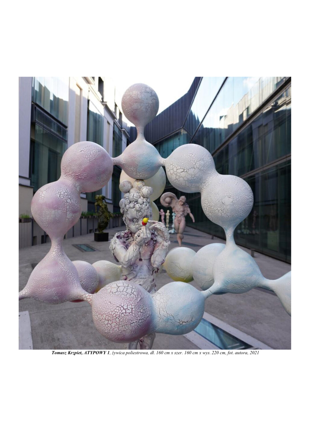 Rzeźba chłopca zbudowanego z atomów, który stoi na kulistym molekule i otoczony jest również molekułami. Kompozycja rzeźbiarska w kolorystyce barw podstawowych (żółtym, czerwonym i niebieskim). Rzeźba jest widoczna w zbliżeniu od głowy do pasa.