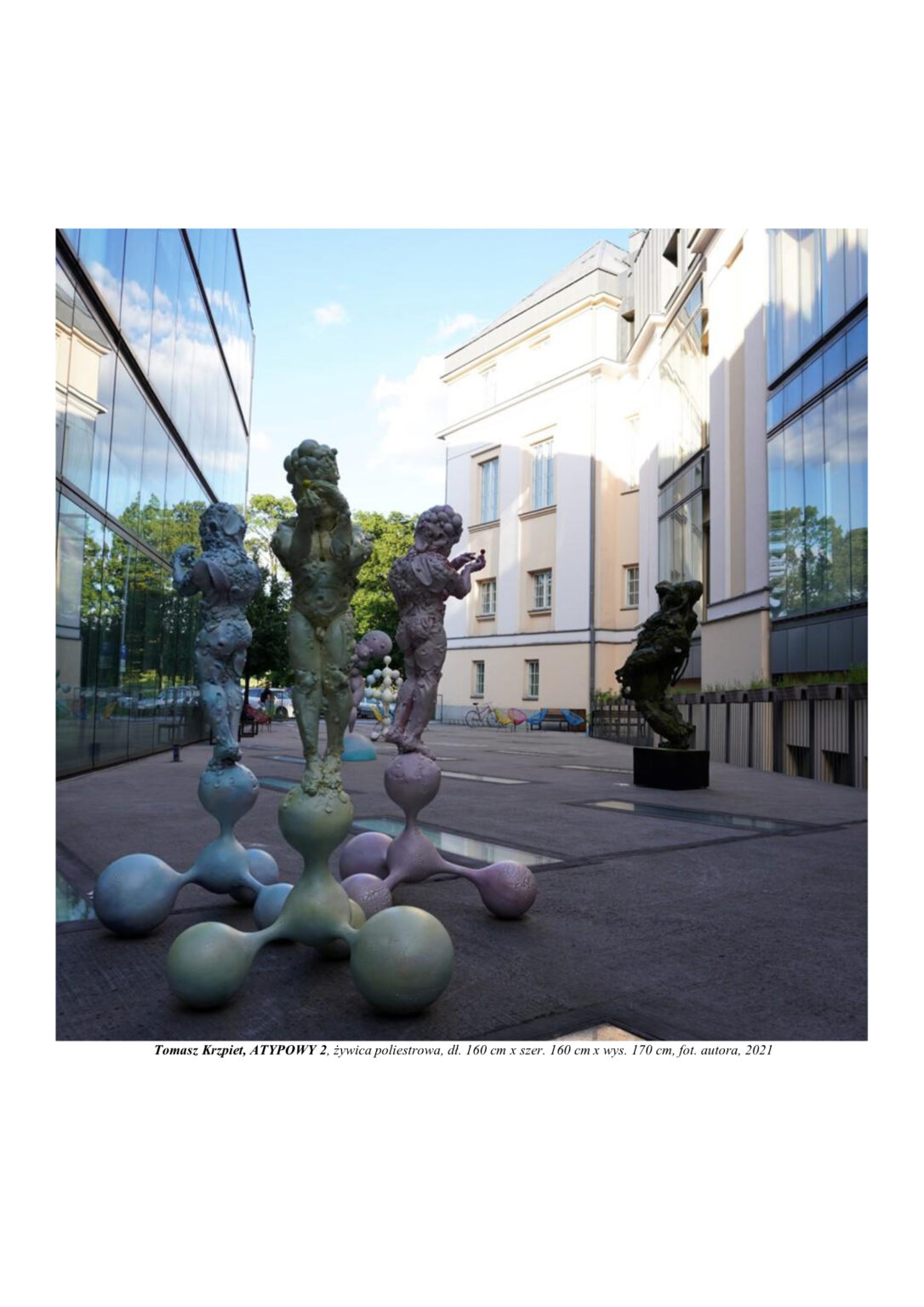 Rzeźba trzech chłopców zbudowanych z atomów, którzy stoją odwróceni plecami od siebie na kulistych molekułach. Kompozycja rzeźbiarska w kolorystyce barw podstawowych (żółtym, czerwonym i niebieskim)