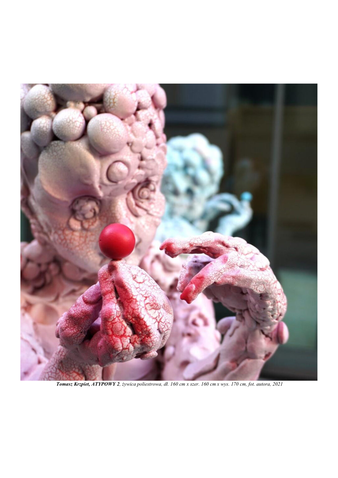 Rzeźba chłopca zbudowanego z atomów, który w palcach trzyma trzy atomy w kolorach żółtym, czerwonym i niebieskim. Kompozycja rzeźbiarska w kolorystyce barw podstawowych (żółtym, czerwonym i niebieskim). Rzeźba jest widoczna w zbliżeniu od głowy do pasa.